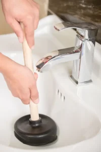 Person using a plunger in a bathroom sink next to a chrome faucet.