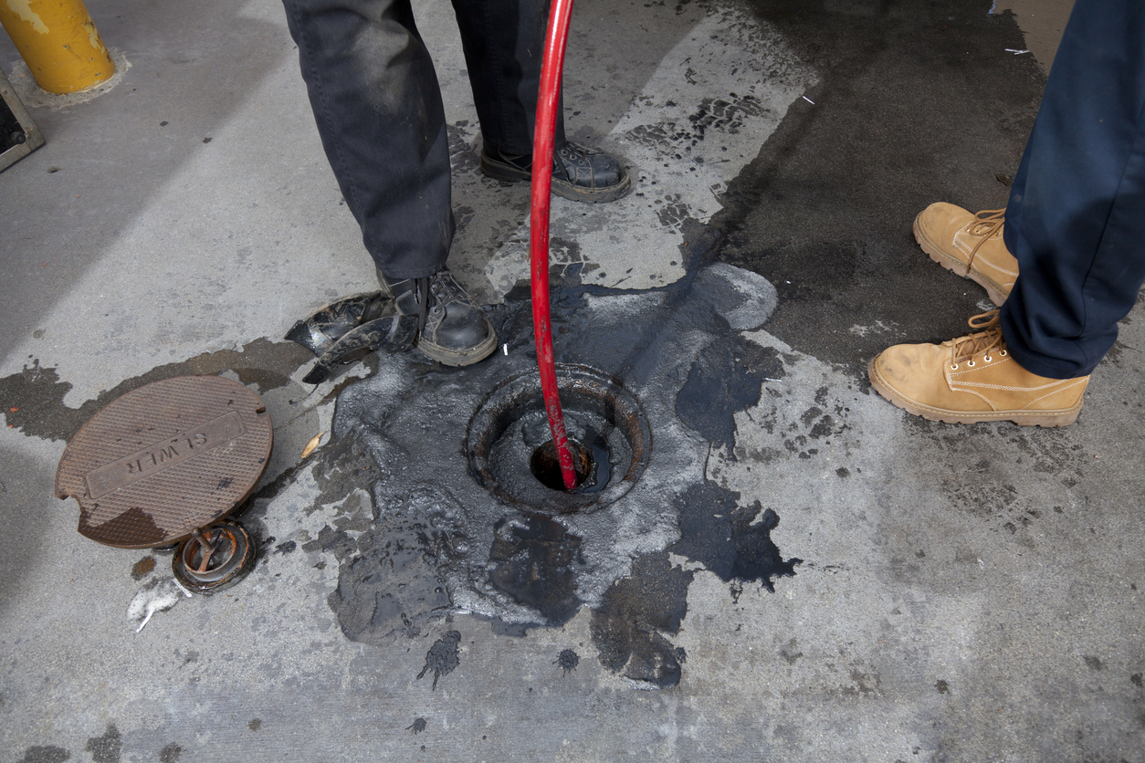 plumbers hydro line jet a pipe at a large restaurant. Grease can be seen on the pavement surrounding the pipe.